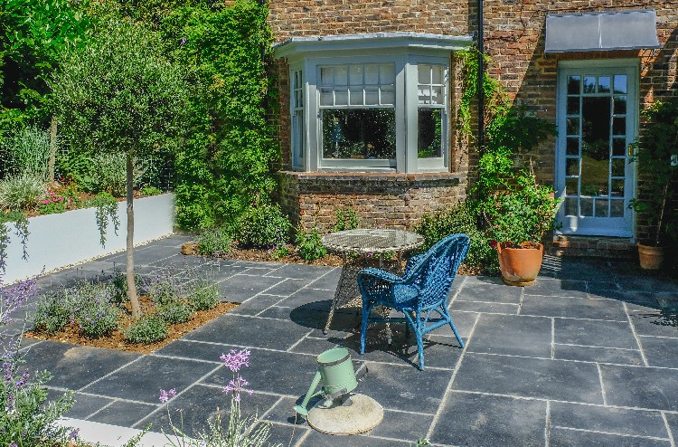 Blue wicker chair and wicker table in the middle of a slate patio with plants and trees in beds and pots.   Taken on a sunny summer day with nobody in the shot.